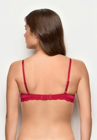 Kobieta z długimi brązowymi włosami widziana od tyłu, ubrana w czerwoną biustonosz z regulowanymi ramiączkami i koronkowymi detalami na jednolitym jasnym tle.