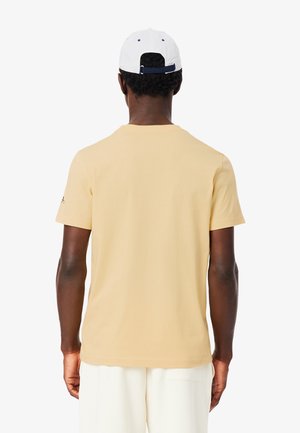 Homme portant un t-shirt beige à manches courtes, un pantalon blanc et une casquette de baseball blanche, vu de dos sur un fond uni.