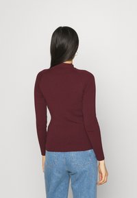 Top lungo a maniche lunghe bordeaux con collo alto e taglio aderente, abbinato a jeans a vita alta blu chiaro.