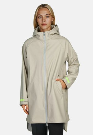 Junge Frau trägt einen hellbeigen Regenmantel mit Kapuze, Frontreißverschluss und neongrünen Akzenten an den Bündchen, steht mit einer Hand in der Tasche.