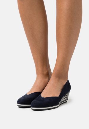 Jambes portant des chaussures à talons compensés en daim bleu marine avec un talon à motifs et des semelles noires, sur un fond blanc.