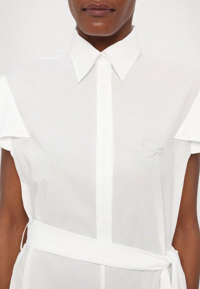 White collared shirt with short sleeves, featuring a front button placket and a waist tie. Subtle metallic logo embroidery on the chest.