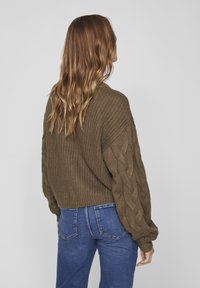 Kvinna med långt, vågigt hår som bär en brun grovt stickad tröja med kabelstickade ärmar och blå denimjeans, vänd bort mot en grå bakgrund.