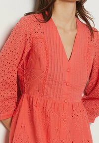 Robe corail avec des manches en broderie anglaise, des plis verticaux et des motifs floraux brodés. Fermeture à boutons et jupe froncée.