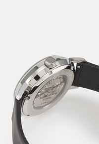 Montre en acier inoxydable avec un fond de boîte transparent, des accents argentés et un bracelet en silicone noir. Dispose d'une couronne pour régler l'heure.