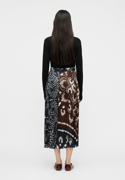 Γυναίκα με μακριά σκούρα μαλλιά φοράει μαύρο μακρυμάνικο τοπ, πλισέ μίντι φούστα με μπλε και καφέ λουλουδένια σχέδια, και σκούρα καφέ παπούτσια.