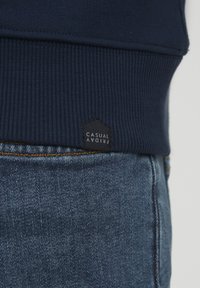 Hanorac navy cu tiv elastic și etichetă din material inscripționată "CASUAL FRIDAY", asortat cu blugi luați cu cusături vizibile.