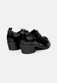 Zapatos de cuero negro con punta redondeada y tacón bajo cuadrado. Presenta un suave detalle plisado y un logo grabado en la parte trasera.