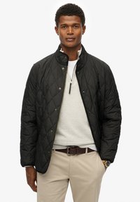 Sort quiltet jakke med høj krave, knappet foran og guld detaljer, parret med en lys grå sweater og beige bukser.