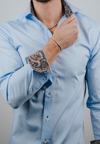 Man die de kraag van een lichtblauw overhemd met paisley-patroon aan de binnenkant van kraag en manchetten rechtzet, met een zilveren armband en ketting aan.