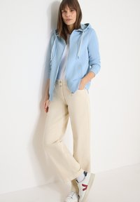 Femme debout contre un mur blanc, portant un sweat à capuche bleu clair, un t-shirt blanc, un pantalon large beige et des baskets blanches avec un détail bordeaux.