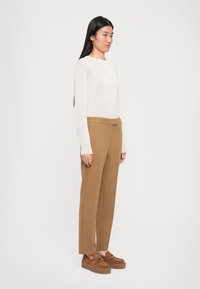 Une femme se tient de profil, portant un pull blanc à manches longues, un pantalon camel et des mocassins compensés marron, devant un fond blanc uni.