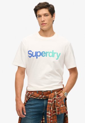 Witte katoenen t-shirt met een blauw-groene gradient "Superdry" logo. Draagt een blauwe spijkerbroek en een oranje geruiten shirt om de taille gebonden.