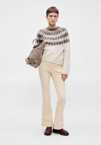 Πλεκτό πουλόβερ με πολύχρωμο σχέδιο στον γιακά, συνδυασμένο με μπεζ φαρδιά παντελόνια και καφέ δερμάτινα παπούτσια. Ένα γκρι υφασμάτινο τσάντα κρατιέται με το ένα χέρι.