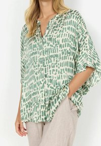 Blouse à motifs verts et crème avec fermeture à boutons sur le devant, coupe décontractée et manches chauve-souris, associée à un pantalon beige clair.