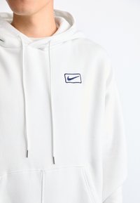 Sweatshirt branco com um grande bolso frontal, cordões com pontas de metal e um pequeno logótipo da Nike bordado em azul no peito.