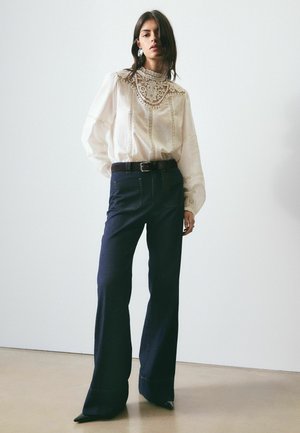 Nő fehér csipkés blúzt visel, ami be van tűrve magas derekú, sötét, széles szárú farmernadrágba fekete övvel, és egy egyszerű fal előtt áll.