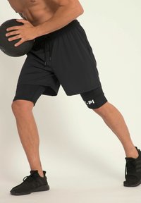 Sorte atletiske shorts med integrerede kompressionsshorts, elastisk talje og logodetalje. Modellen holder en medicinbold.