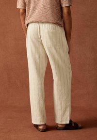 Lys beige bukser i en struktureret stof har lige ben, elastisk talje og en enkelt baglomme. Styling med mørke sandaler.