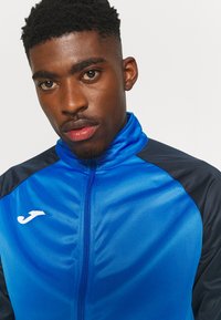 Giacca sportiva blu con rifiniture blu navy, chiusura a zip frontale, colletto alto e una leggera lucentezza. Presenta un logo bianco sul lato sinistro del petto.