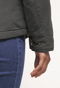 Main droite d'une personne portant une veste vert foncé avec un poignet boutonné et un jean en denim bleu, sur un fond neutre.