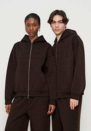 Due modelli che indossano giacche con cappuccio marrone scuro abbinate con zip e pantaloni, in piedi vicini contro uno sfondo chiaro e uniforme.