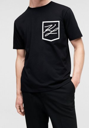 Mies, kalsarivärinen musta lyhythihainen t-paita, jossa on valkoisella ääriviivalla oleva rintatasku, joka esittelee tyyliteltyjä valkoisia alkukirjaimia, yhdistettynä mustiin housuihin.