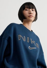 Sötétkék Nike pulóver laza szabással, nagy, krémszínű hímzett logóval, amelyen egy swoosh látható. Puha, texturált pamut anyagból kész ült.