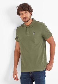 Man with curly hair wearing an olive green polo shirt and blue jeans standing against a white wall.