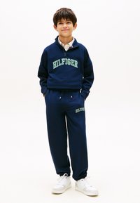 Αγόρι που στέκεται με τα χέρια στις τσέπες, φορώντας σκούρο μπλε φόρμα Hilfiger και λευκά αθλητικά παπούτσια, μπροστά σε ένα απλό λευκό φόντο.
