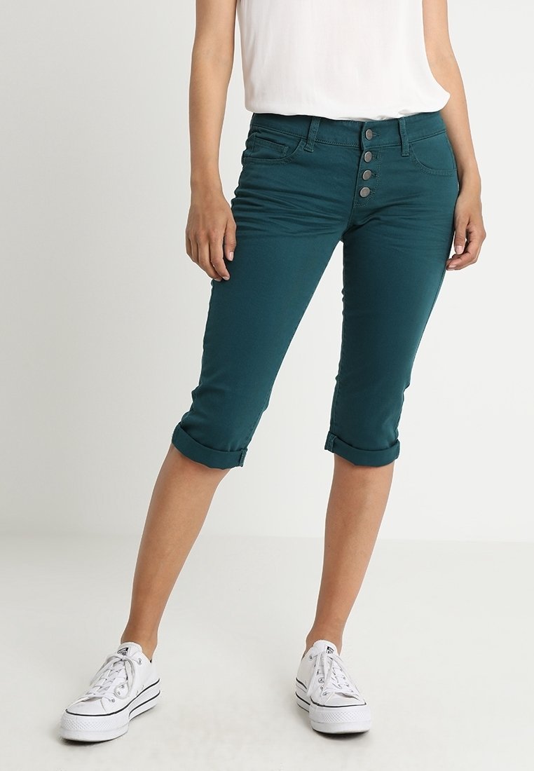 Femme portant un pantacourt bleu sarcelle foncé retroussé avec une braguette à boutons, associé à des baskets blanches et un haut sans manches blanc.
