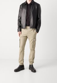 Černá kožená bunda, upnuté černé polo tričko, béžové cargo kalhoty s bočními kapsami a černé kotníkové boty. Hladké textury a ležérní design.