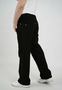 Svarta denimbyxor med rak passform, fem fickor och kopparfärgade nitar, tillsammans med vita sneakers, visade från baksidan.