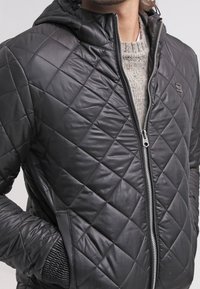 Czarna pikowana kurtka z kapturem, z zamkiem z przodu, bocznymi kieszeniami i teksturowanymi mankietami. Wykonana z materiału syntetycznego, o gładkim wykończeniu.