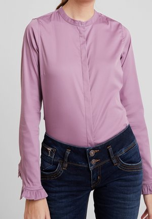 Kvinde iført en langærmet lavendelbluse med flæser ved manchetterne og mørkeblå højtaljede jeans med dobbelte knapper foran og kontrastsyninger.