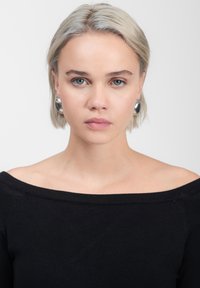 Maglione nero a spalle scoperte, dal tessuto morbido; modella con capelli corti e biondi chiari, grandi orecchini d'argento e trucco naturale. Sfondo neutro.