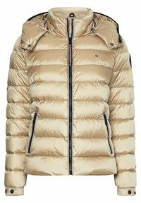 Beige vadderad jacka med hög krage, dragkedjestängning och två sidofickor. Har en huva och kontrasterande svarta detaljer. Slät textur.