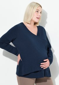 Marineblaue, langärmelige Umstand bluse mit V-Ausschnitt und asymmetrischem Saum. Weicher Stoff mit lockerer Passform, getragen mit beigen Leggings.