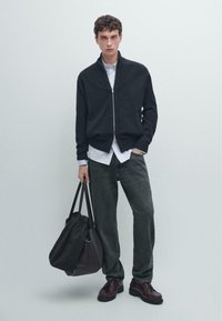 Maglione zip-up scuro, indossato sopra una camicia bianca con bottoni, abbinato a jeans scuri. Scarpe nere e una grande borsa grigia con manici neri.