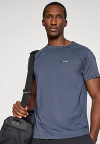 Ναυτικό μπλε αθλητικό μπλουζάκι με κοντά μανίκια, σχέδιο ραγλάν, ύφασμα που απομακρύνει την υγρασία και μικρό λογότυπο στο στήθος, σε συνδυασμό με μία μαύρη τσάντα.