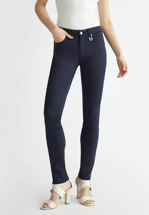 Femme portant un jean skinny foncé avec un haut sans manches côtelé blanc et des sandales à talons hauts crème, debout avec une main dans la poche.