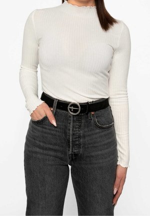 Femme portant un haut blanc côtelé à manches longues, rentré dans un jean en denim noir taille haute, accompagné d'une ceinture noire avec une boucle circulaire argentée.