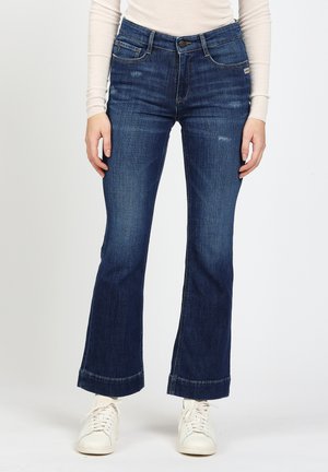Person wearing dark blue slightly flared jeans with subtle distressed details, a beige long-sleeve top, and white sneakers.