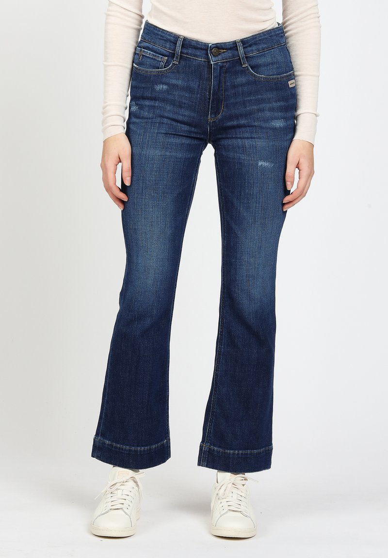Person wearing dark blue slightly flared jeans with subtle distressed details, a beige long-sleeve top, and white sneakers.