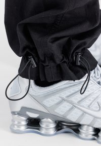 Primer plano de una zapatilla deportiva blanca y plateada con una suela tipo resorte, llevada con pantalones negros ajustados por cuerdas de ajuste.