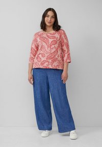 Rotes Top mit Paisleymuster und Dreiviertelärmeln, kombiniert mit blauen weiten Hosen und weißen Sneakers. Glatte Stoffstruktur.