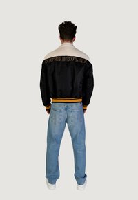 Giacca bomber nera e crema con "SUPER BOWL LVII" ricamato sulla schiena, polsini e orlo a costine, abbinata a jeans blu chiaro.