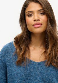 Pull en tissu bleu texturé avec un large décolleté et des manches amples, porté par un mannequin aux cheveux ondulés et au maquillage minimaliste.