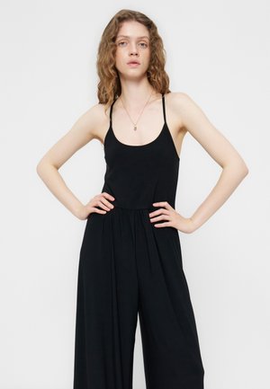 Γυναίκα με σγουρά μαλλιά που φοράει μαύρη αμάνικη φόρμα με λεπτές τιράντες και έχει τα χέρια στη μέση, με φόντο ένα απλό ανοιχτόχρωμο φόντο.