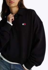 Zwarte oversized polo sweatshirt met een geribde kraag en manchetten, voorzien van een klein 'Tommy Jeans' logo op de linkerkant.